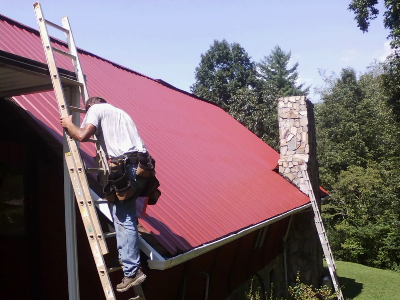 Roofer on a ladder beside a red metal roof and stone chimney — Soffit & Fascia Repair by Roofing Brothers in Mickleton